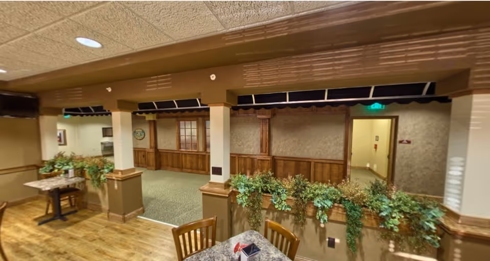 Communal dining area with wooden tables and chairs, indoor plants, and wood-paneled walls under a drop ceiling.