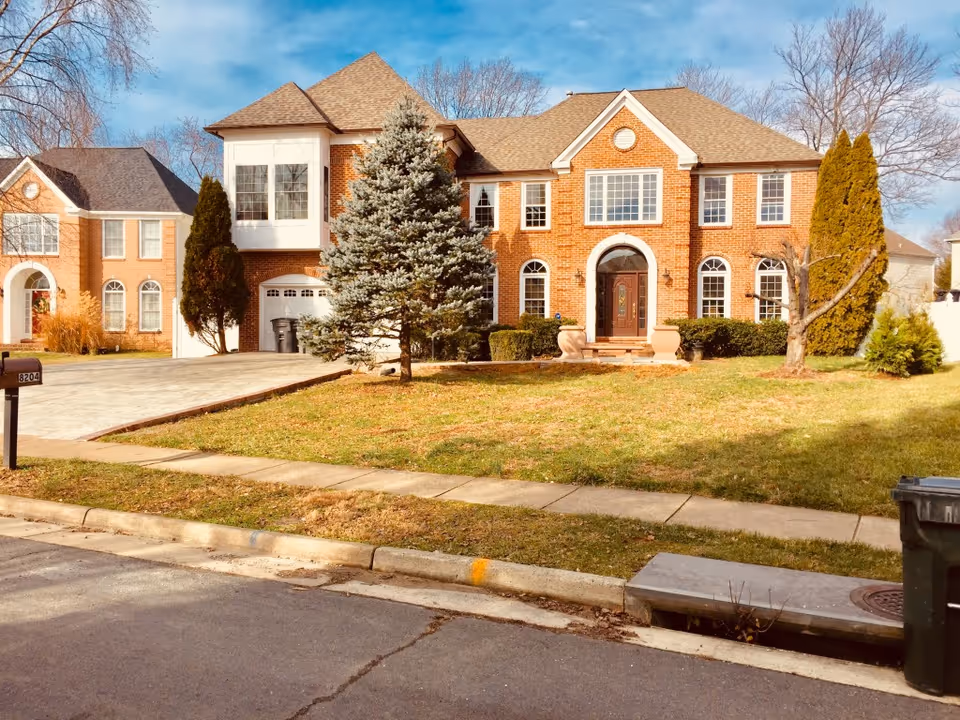 Front exterior view of a large two-story brick house with multiple windows, a central arched doorway, a driveway, and a lawn with trees and shrubs under a blue sky.