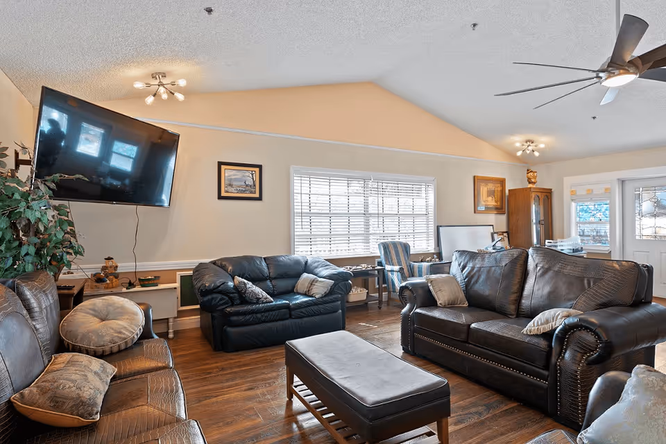 A cozy living room with multiple leather sofas and armchairs arranged around a central ottoman. A large flat-screen TV is mounted on the wall to the left. The room has wooden flooring, a ceiling fan, and several framed pictures on the walls. A large window with blinds lets in natural light, and there is a cabinet and a door with decorative glass on the right side.