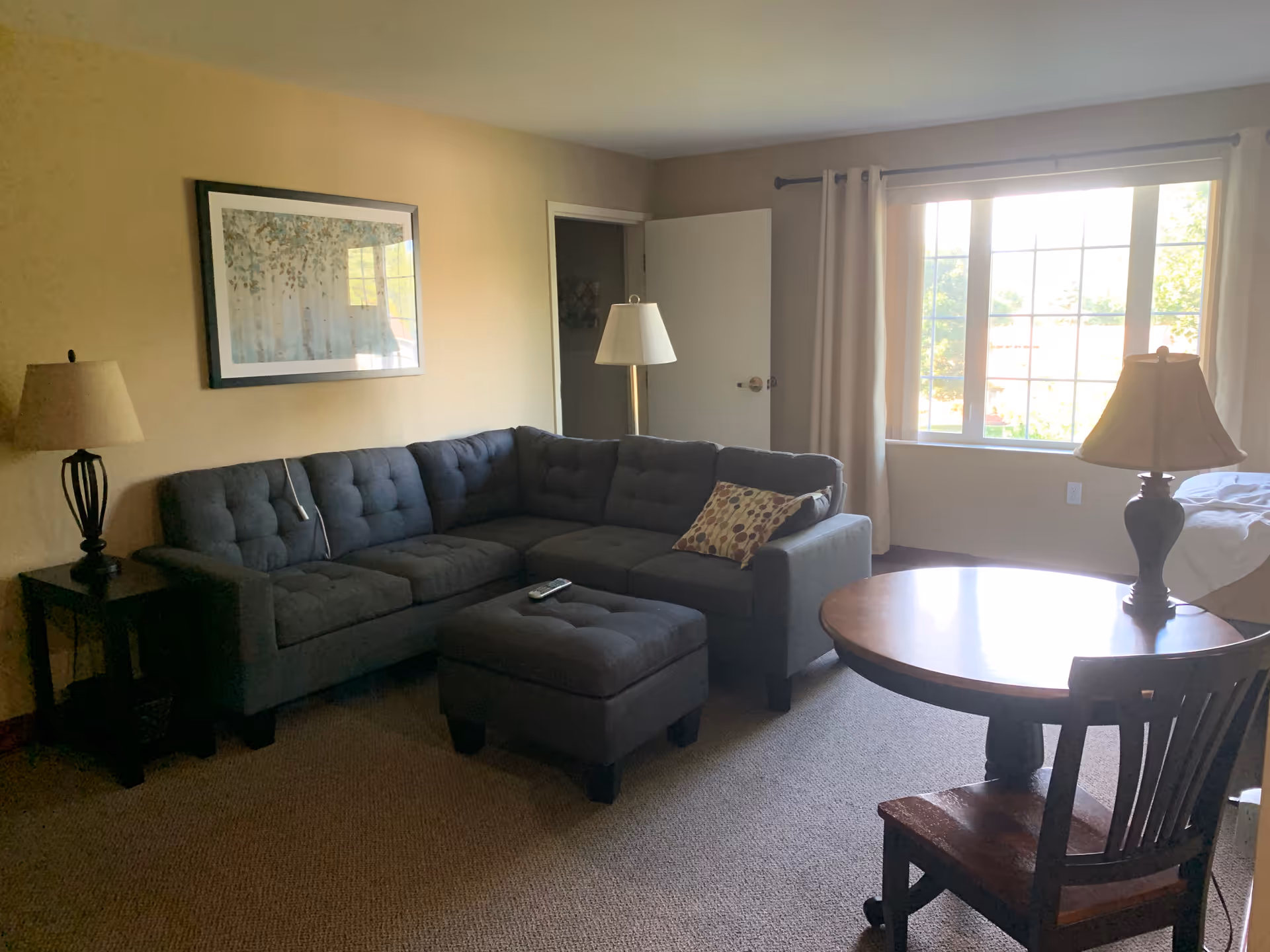 A cozy living room with a dark gray sectional sofa and matching ottoman, a wooden side table with a lamp, a round wooden table with a lamp and chair, a large window with beige curtains letting in natural light, and a framed artwork on the beige wall.