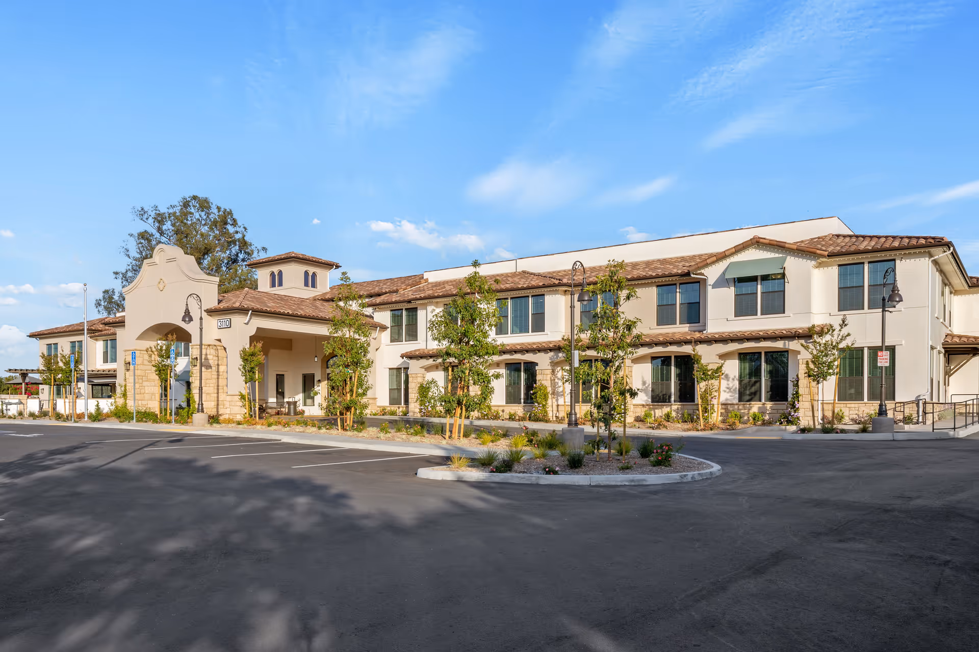 Exterior view of a two-story senior living facility building with beige walls and a tiled roof under a clear blue sky. The building has multiple windows, a covered entrance with stone pillars, and a parking lot with some landscaping and young trees in front.