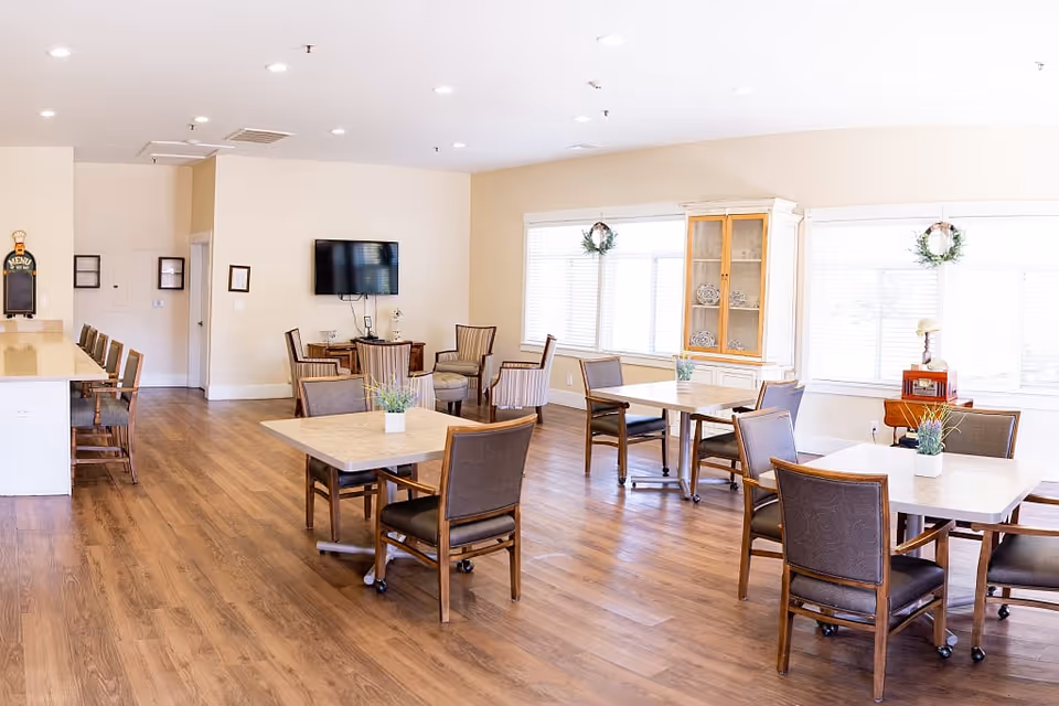 A bright and spacious senior living dining area with wooden flooring, several square tables each surrounded by four chairs, large windows with white blinds, a wall-mounted TV, and a cabinet displaying decorative items. There is also a counter with high chairs on the left side and some small plants on the tables.