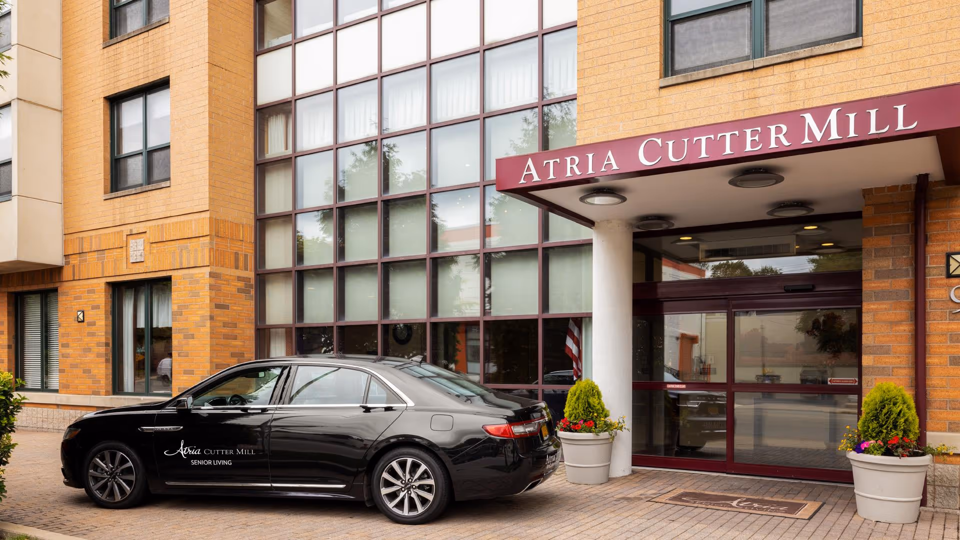 Exterior view of the entrance to Atria Cutter Mill senior living facility, featuring a maroon awning with the facility name, large windows, and a black car parked in front with the Atria Cutter Mill logo on its door. Two large potted plants flank the entrance.