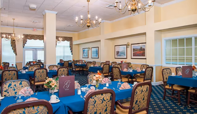 A dining room with multiple tables covered in blue tablecloths, each set with pink folded napkins, glassware, and floral centerpieces. The chairs have a floral patterned upholstery. The room has beige walls with framed artwork and brass chandeliers hanging from the ceiling.