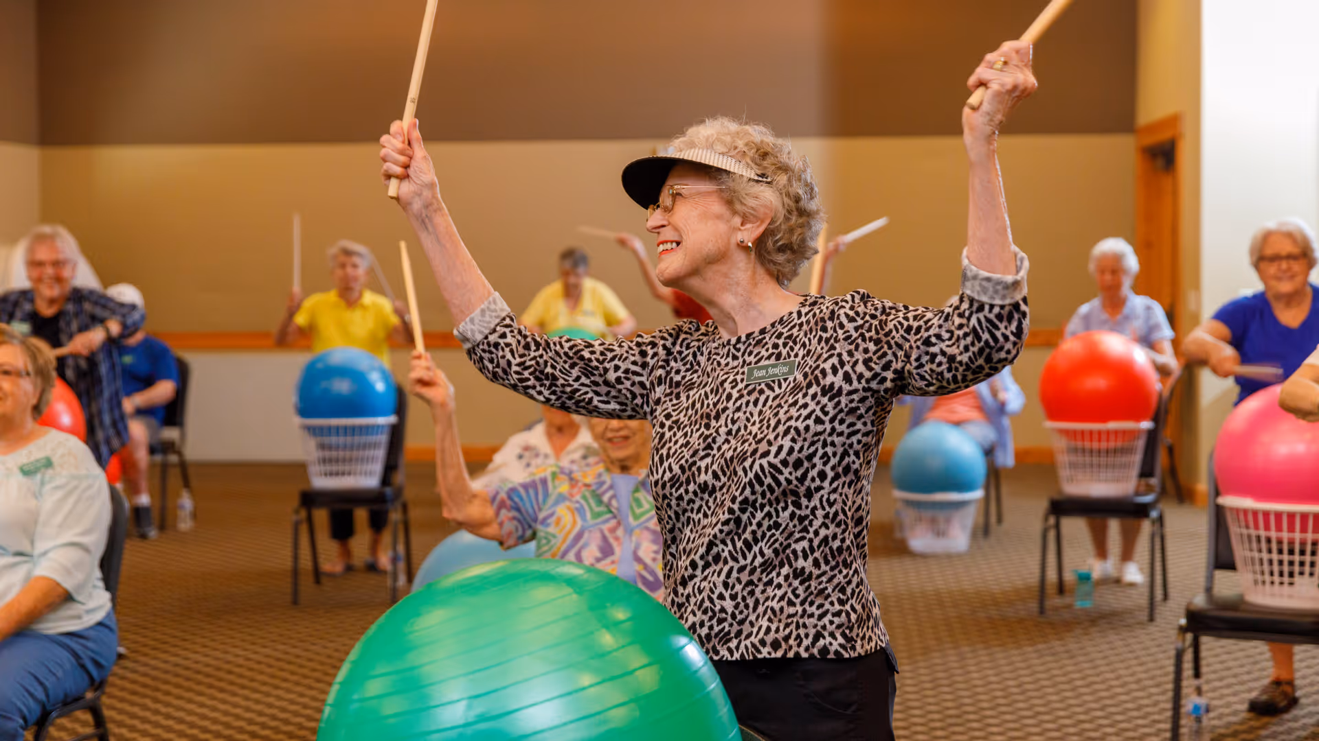 A group of elderly women participating in a seated exercise class using large colorful exercise balls and drumsticks in a spacious room. The woman in the foreground is smiling, wearing a leopard print top and a visor, and holding drumsticks above her head.