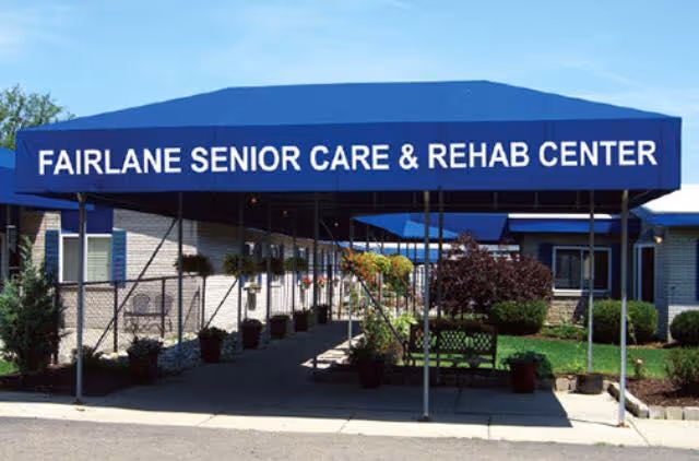 Entrance of Fairlane Senior Care & Rehab Center with a blue canopy displaying the facility name. The walkway is lined with potted plants and greenery on both sides, leading to the building entrance.