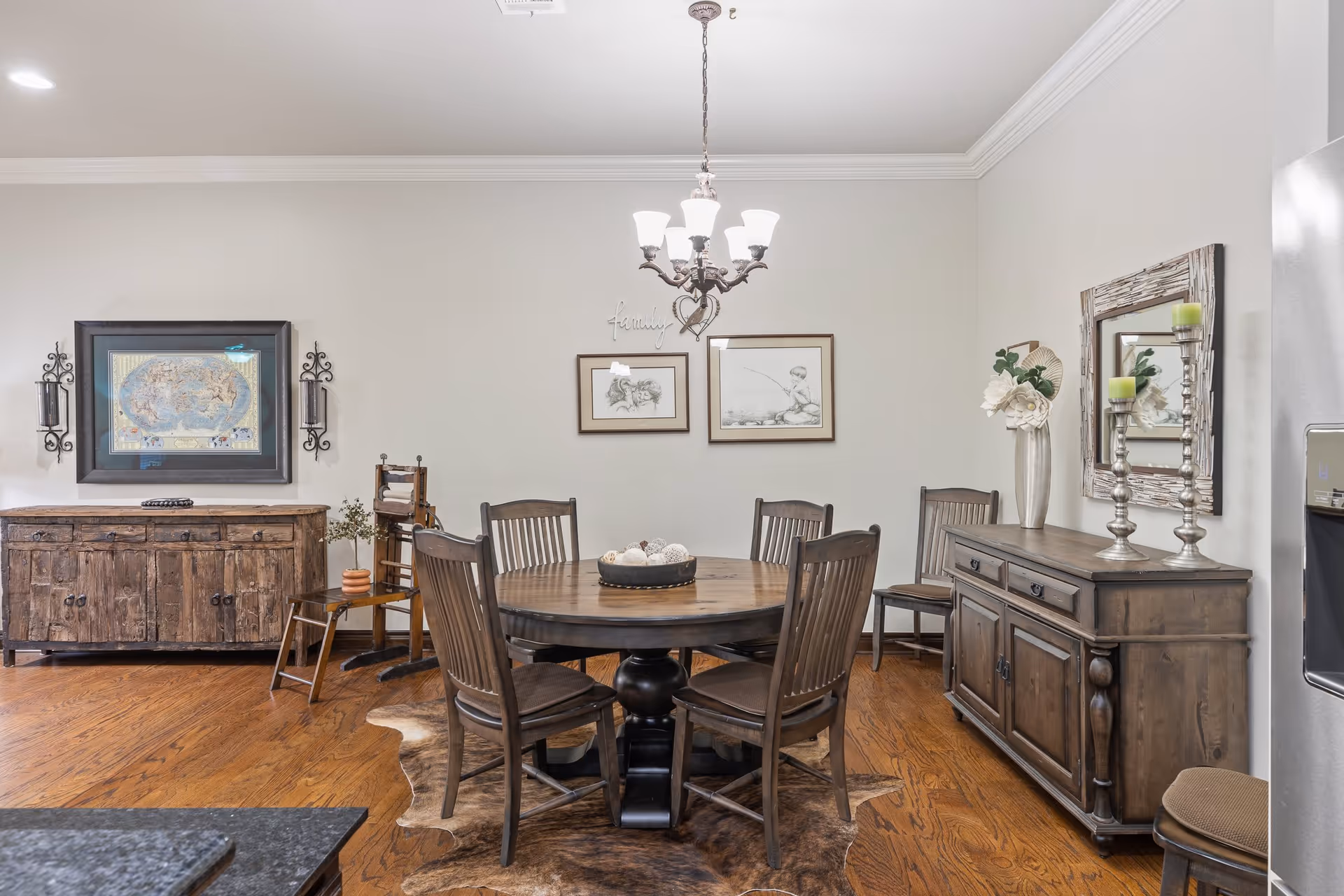 A dining room with a round wooden table surrounded by six wooden chairs. The floor is hardwood with a cowhide rug underneath the table. On the left wall, there is a rustic wooden sideboard with a framed map above it and two wall sconces. On the right wall, there is another wooden sideboard with a mirror above it, decorated with a vase of flowers and two candle holders. Two framed sketches and a chandelier with six lights hang on the wall in the center.