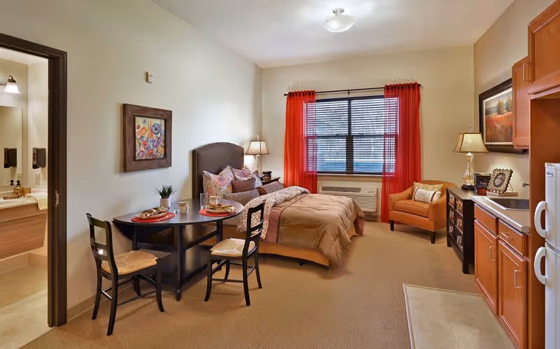 A cozy senior living facility room featuring a bed with multiple pillows and a beige comforter, a small round dining table set for two with plates and glasses, an armchair near a window with red curtains, a kitchenette with wooden cabinets and a microwave, and a bathroom visible through an open door.