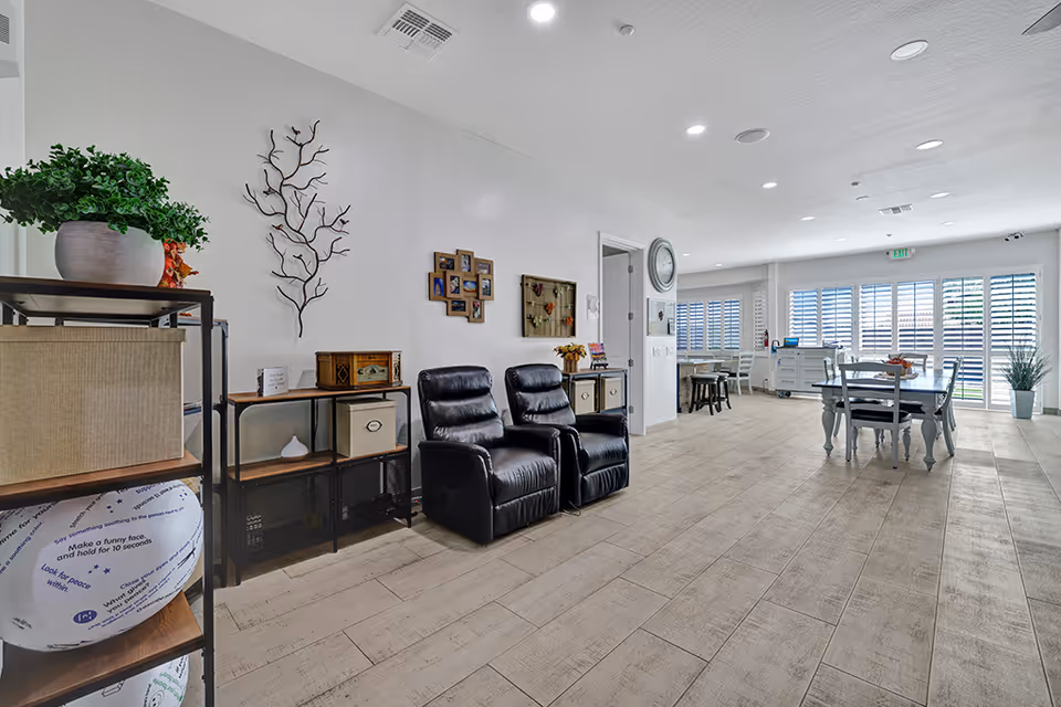 A bright and spacious common area in an assisted living facility featuring two black leather recliners, a dining table with chairs near large windows with white shutters, decorative wall art, and shelves with plants and storage boxes.