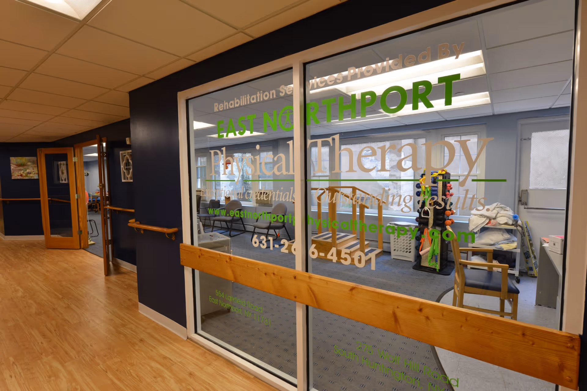 View of a hallway in a senior living facility with wooden handrails on the walls and a glass window showing a physical therapy room. Inside the therapy room, there are exercise equipment, chairs, and a set of stairs for rehabilitation exercises. The glass window has text advertising East Northport Physical Therapy with contact information.