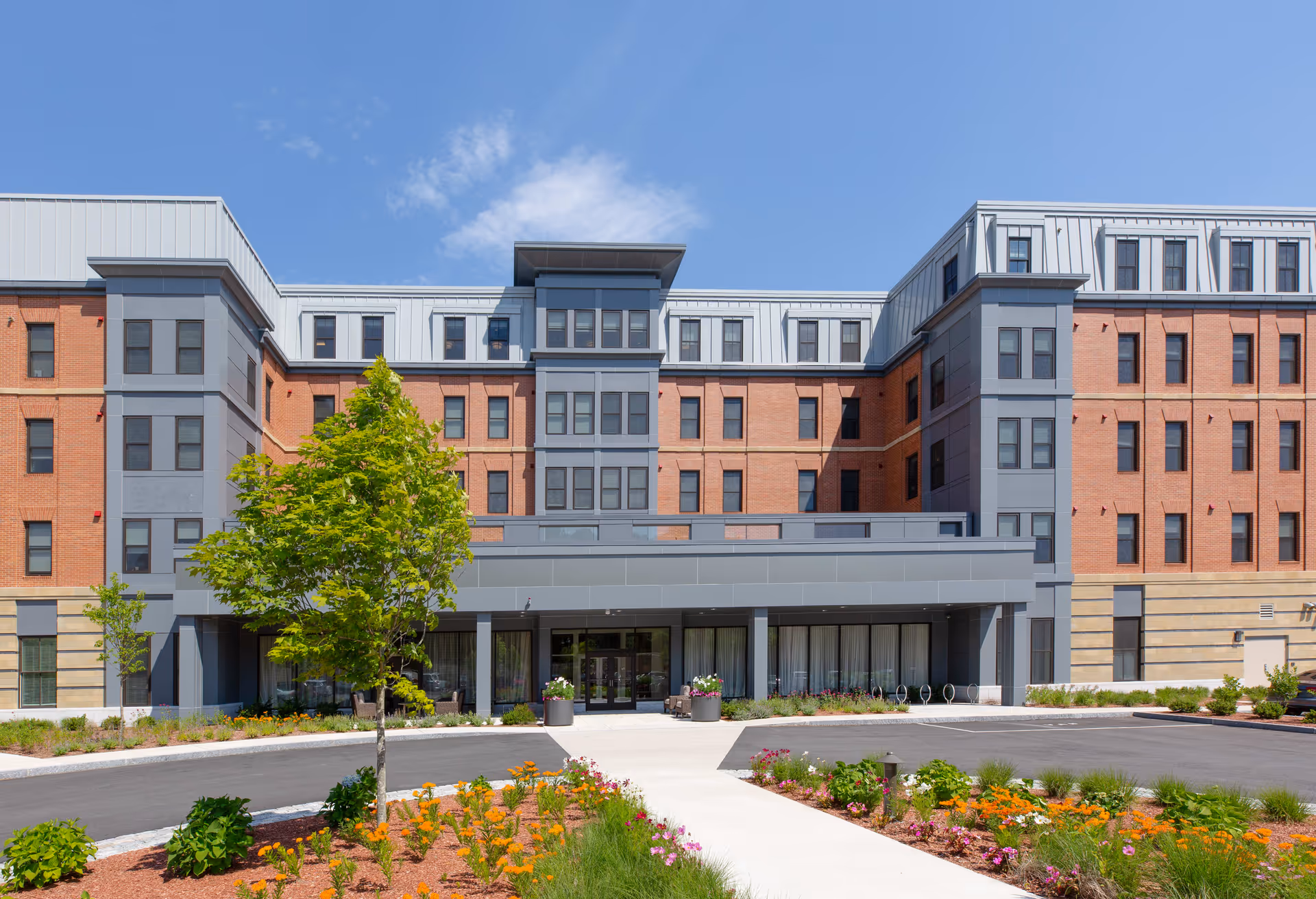 Front exterior view of a multi-story senior living facility with a brick and gray facade, large windows, a covered entrance, and landscaped flower beds with a tree and colorful flowers under a clear blue sky.