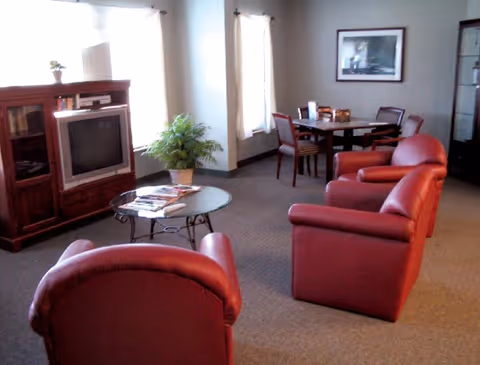 A living room area with three red leather armchairs arranged around a glass coffee table with magazines on it. There is a wooden TV cabinet with an older-style television, a potted plant, and a dining table with four chairs in the background near two windows with curtains. A framed picture hangs on the wall.