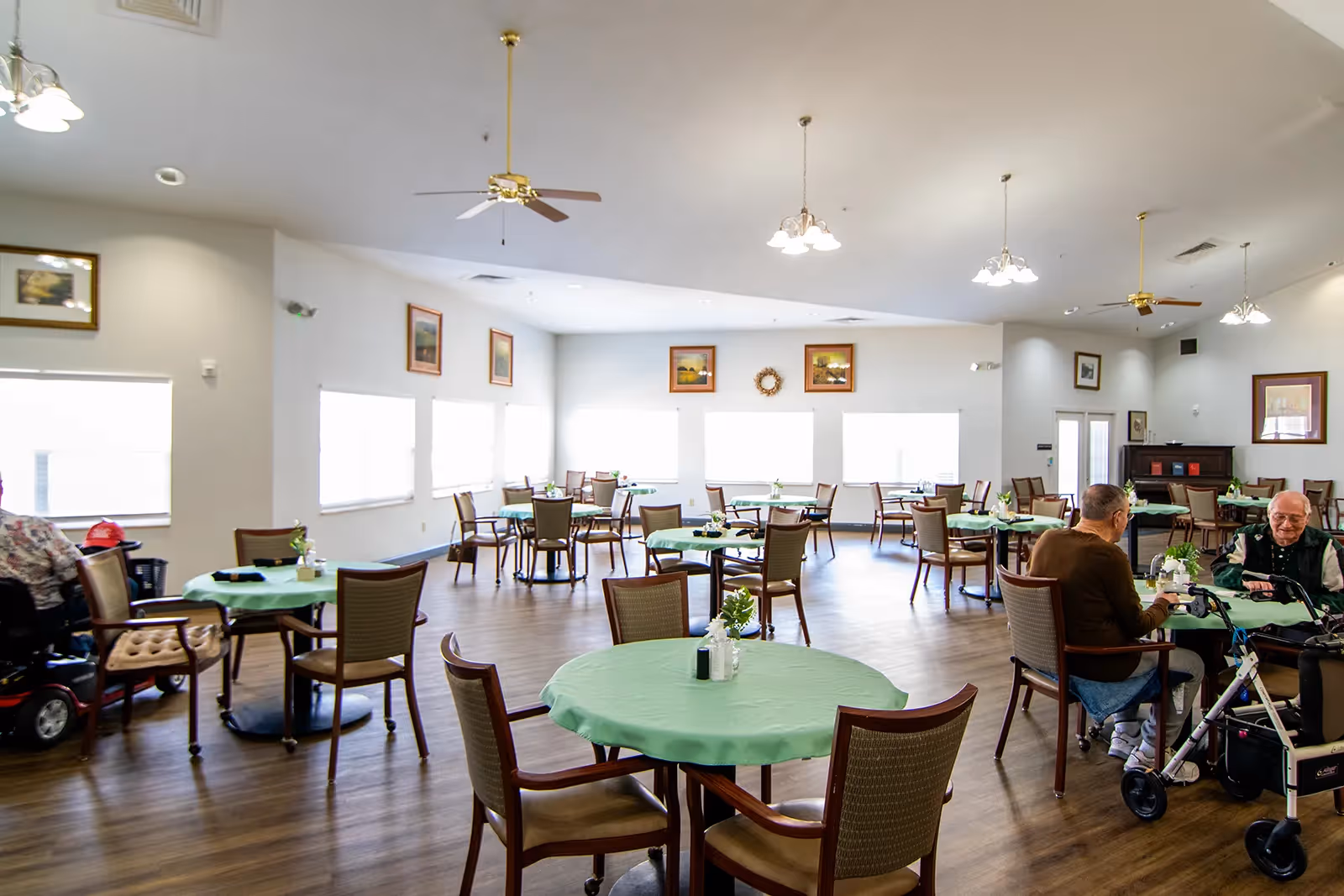 A spacious dining room with multiple round tables covered with green tablecloths, each surrounded by chairs. Two elderly men are seated at one table, engaged in conversation, with one using a walker. The room has large windows letting in natural light, ceiling fans, and framed artwork on the walls.