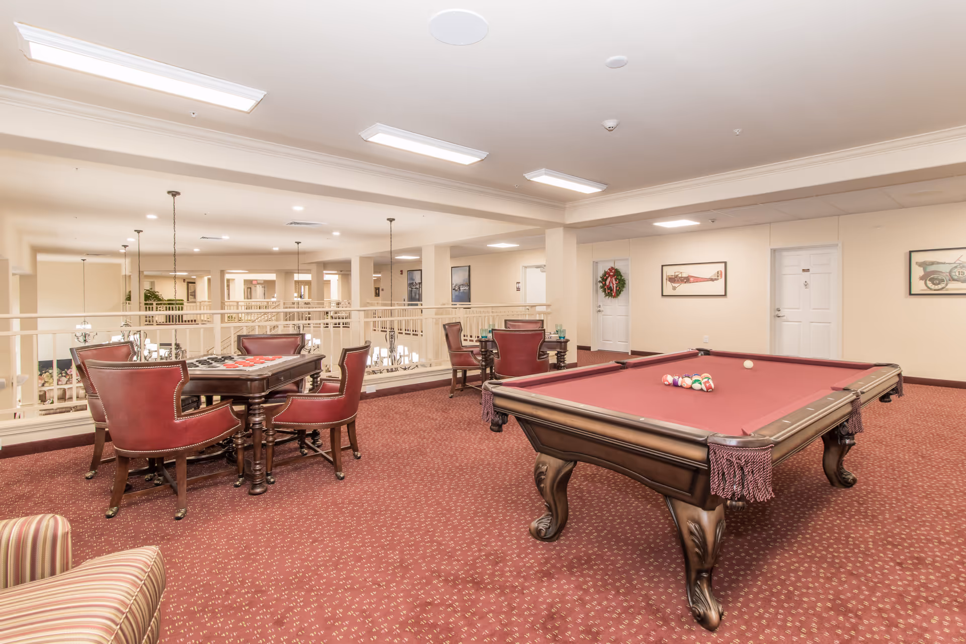 A spacious recreational room with a red carpet featuring a pool table with balls arranged for a game and a card table with four red leather chairs. The room has beige walls adorned with framed pictures and a holiday wreath on one door. Overhead fluorescent lights illuminate the area, and a railing overlooks a lower level.
