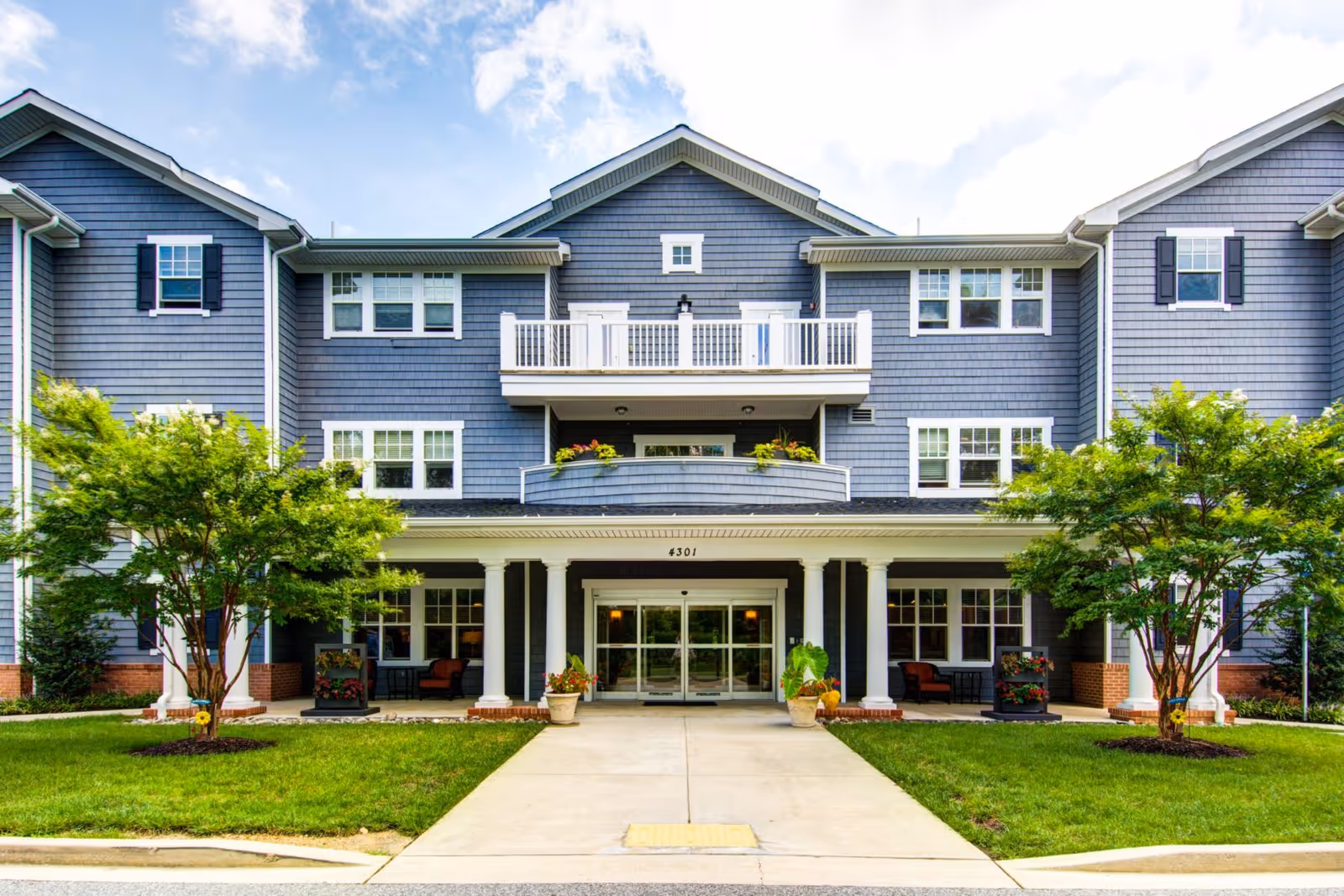 Front exterior view of Symphony Manor, a multi-story building with blue siding, white trim, and a covered entrance supported by white columns. There are two trees and green grass on either side of the concrete walkway leading to the entrance.