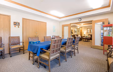A communal room with a long table covered by a blue tablecloth surrounded by eight chairs with patterned upholstery. The room has beige carpet, light-colored walls, and wooden doors. In the background, there is an adjoining room with more seating and a popcorn machine on the right side.