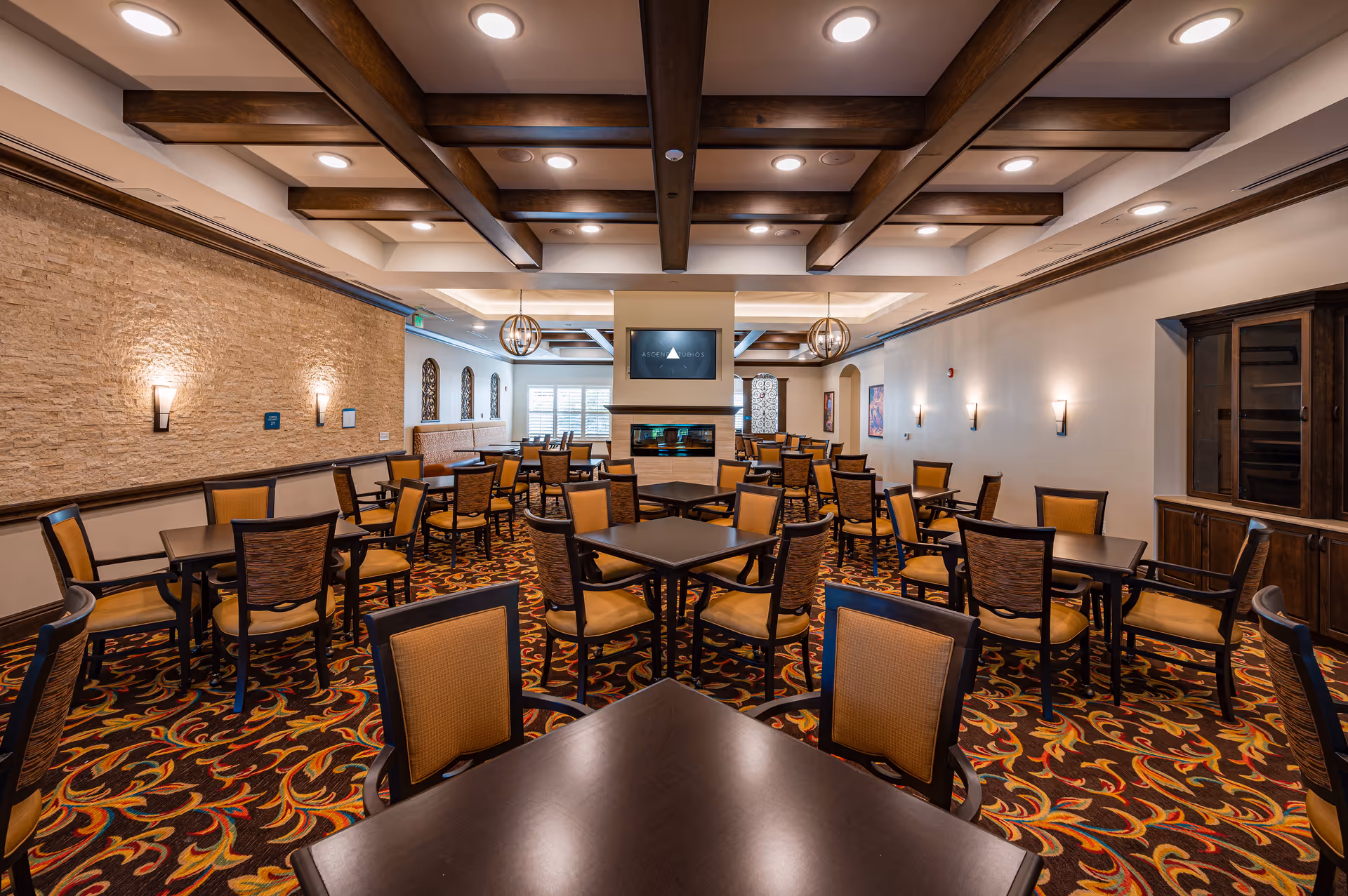 A spacious dining room in Palagio Senior Living with multiple dark wood tables and chairs with tan upholstery. The room features a patterned carpet with red, yellow, and black colors, a stone accent wall with sconces, wooden ceiling beams, and modern pendant lights. A fireplace with a TV mounted above it is visible at the far end of the room.