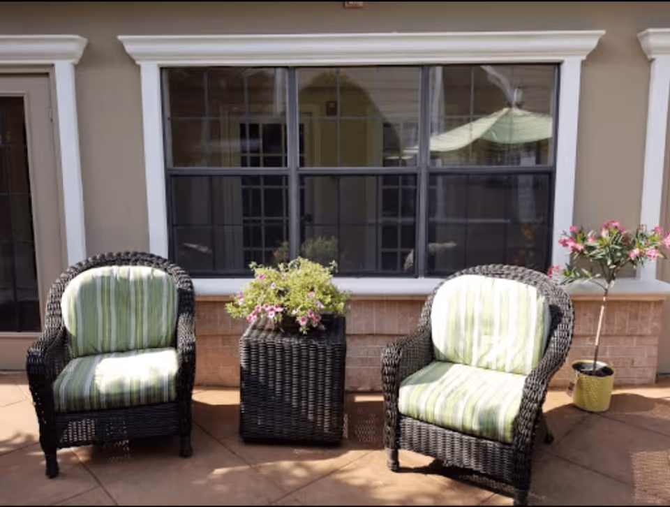 Two wicker chairs with green and white striped cushions placed on a patio in front of a window. Between the chairs is a wicker planter with pink flowers, and to the right is a potted plant with pink flowers. The setting appears to be an outdoor seating area.