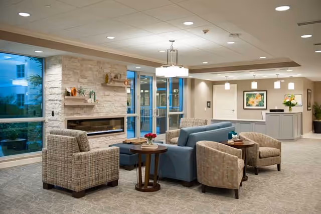 A modern and cozy living room area in a senior living facility featuring a stone fireplace, a blue sofa, several armchairs, side tables with decorative items, and a reception desk in the background. Large windows and glass doors provide natural light and a view of the outdoor area.