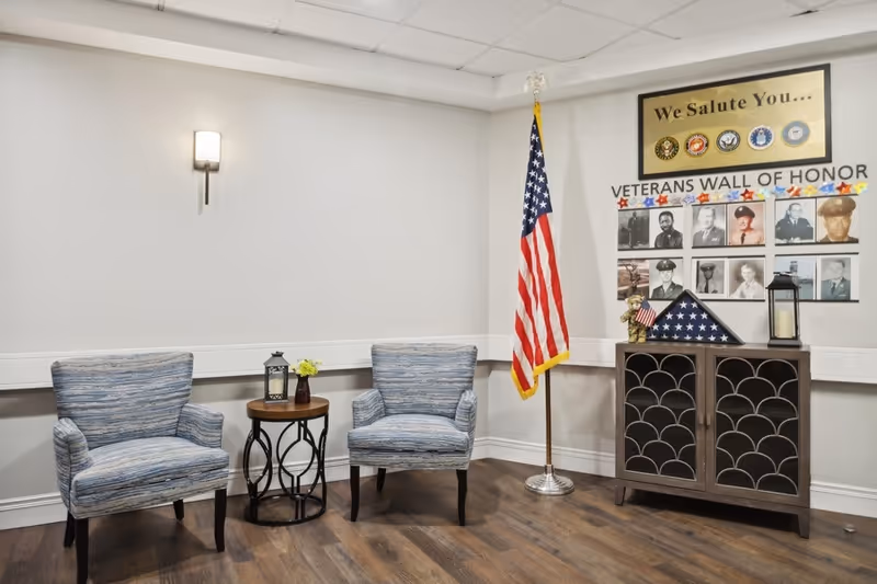 A small seating area with two blue patterned armchairs and a round wooden side table with a lantern and small flower vase on it. To the right, there is a cabinet with a folded American flag, a teddy bear holding a small flag, and a lantern. Above the cabinet is a Veterans Wall of Honor display with photos of veterans and a sign that reads 'We Salute You...' along with military branch emblems. An American flag on a stand is positioned between the chairs and the cabinet. The room has wood flooring and light-colored walls with a wall sconce light fixture.