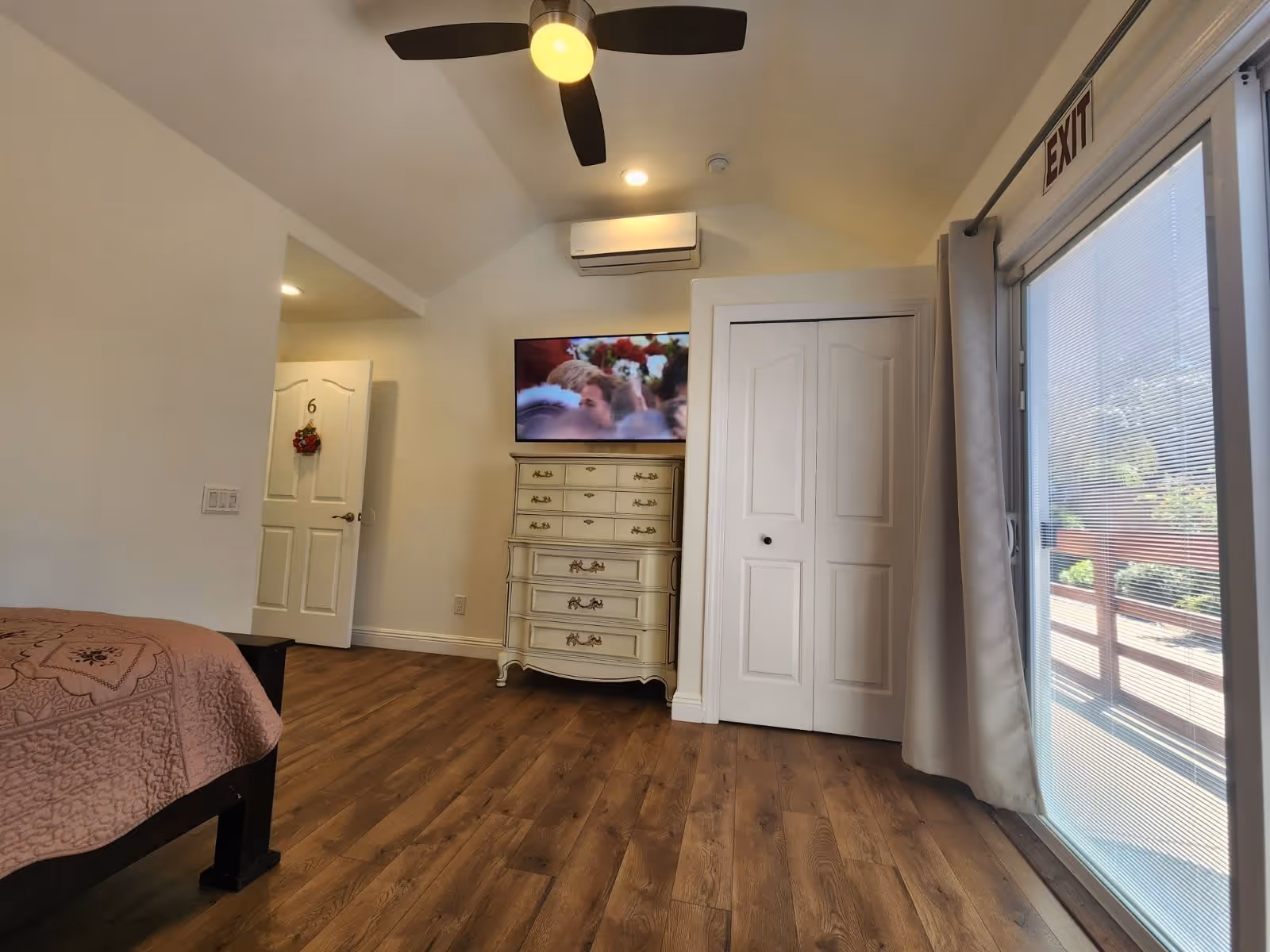 Interior view of a bedroom with wooden flooring, a bed with a pink quilt on the left, a white dresser with a TV mounted above it, a ceiling fan with a light, a door marked with the number 6 slightly open, a closet with double doors, and a glass door with an exit sign leading outside.