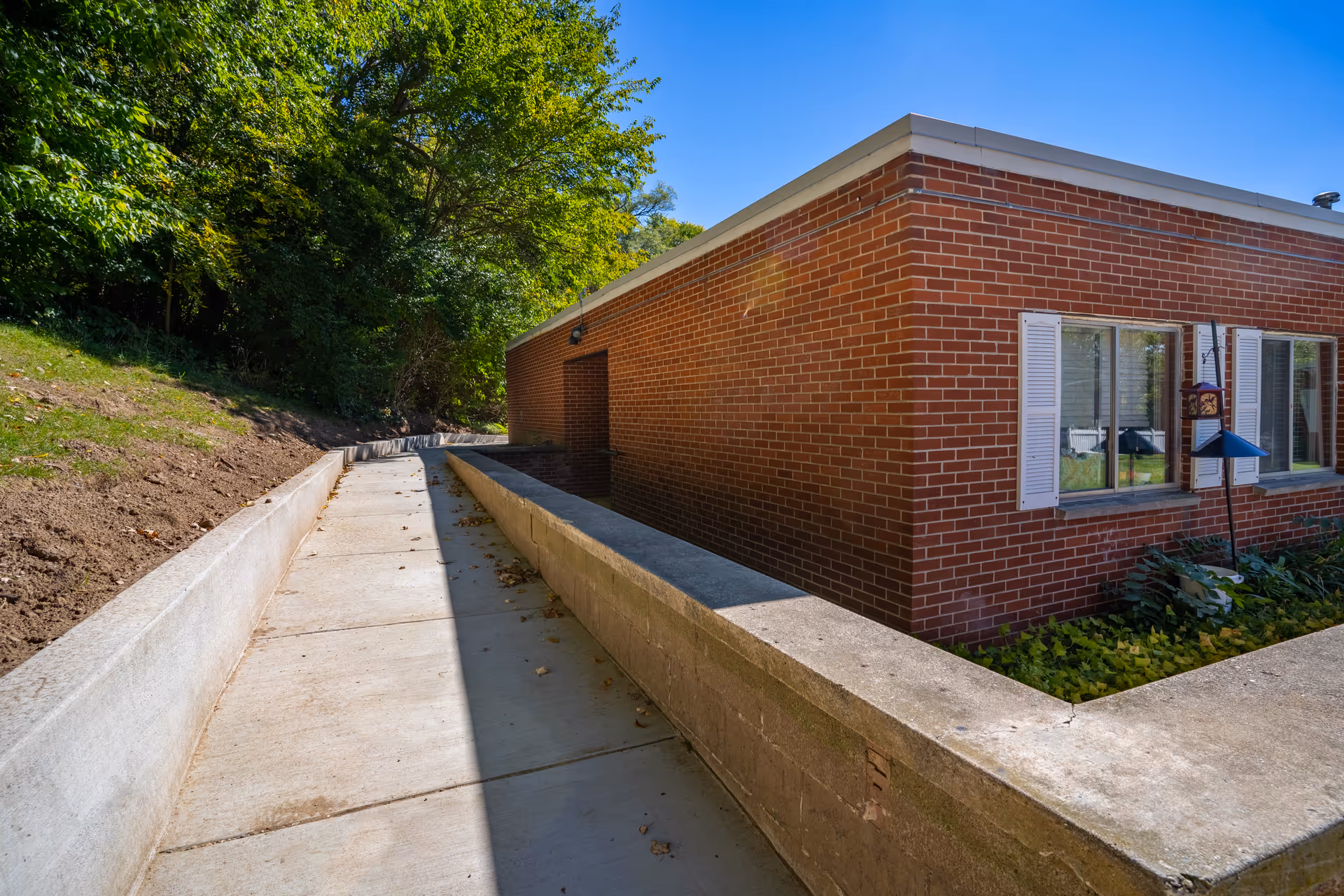Outdoor view of a brick building with white window shutters and a concrete pathway alongside it. The pathway is bordered by a low concrete wall on one side and a dirt slope with greenery on the other. Trees and bushes are visible in the background under a clear blue sky.