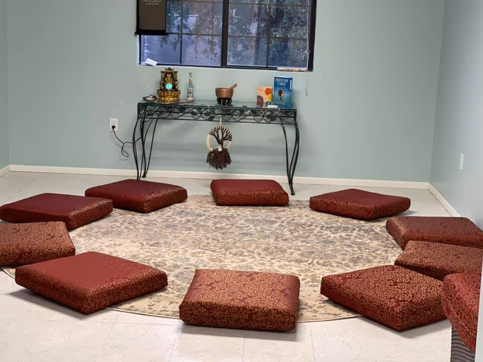 A quiet room with a circular patterned rug on the floor surrounded by red cushions arranged in a circle. Against the wall is a decorative black metal table with various small items on top, including a small statue, a bowl, and some books. A window with closed blinds is above the table.