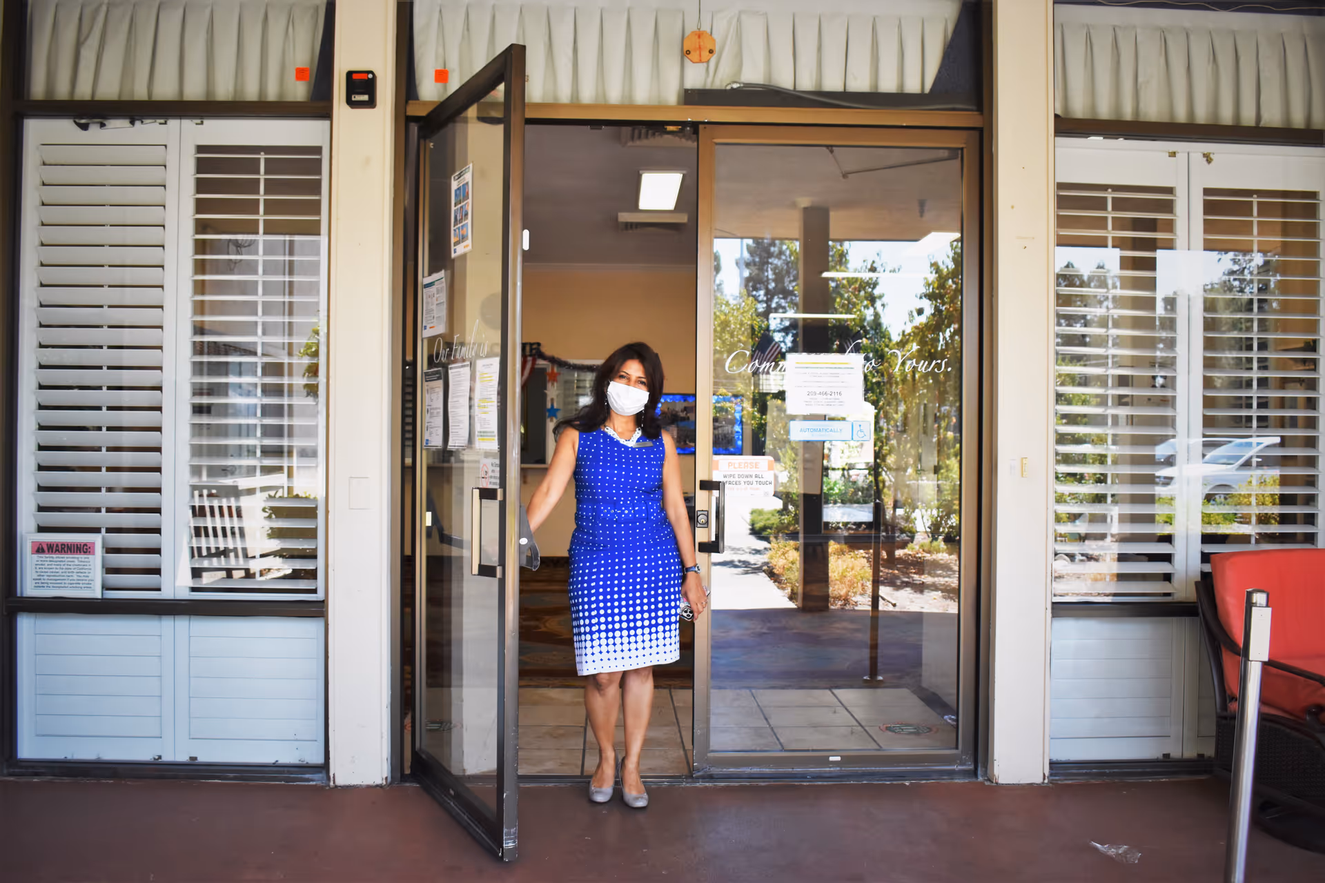 A woman wearing a blue and white dress and a face mask is standing at the entrance door of a building, holding the door open. The entrance has large glass doors and windows with white shutters on either side. There is a red cushioned chair to the right and some signs posted on the glass door.