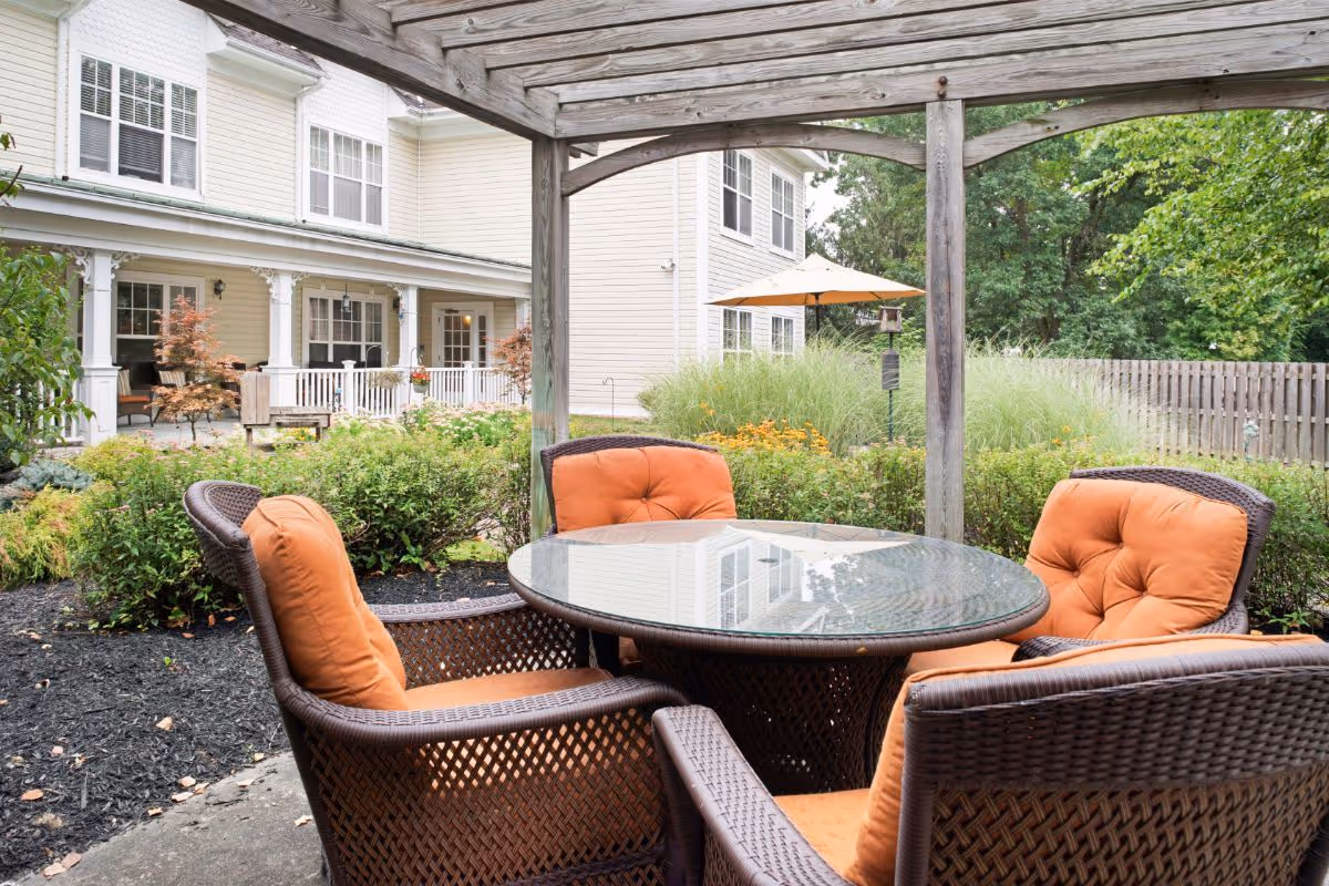 Outdoor courtyard patio with wicker chairs topped with orange cushions around a glass table under a wooden pergola, with the facility building and garden in the background.