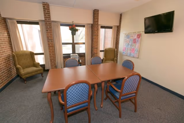 Small communal dining/activity room with a rectangular table surrounded by six chairs, two armchairs by the windows, a wall-mounted TV, and a quilt hanging on the wall.