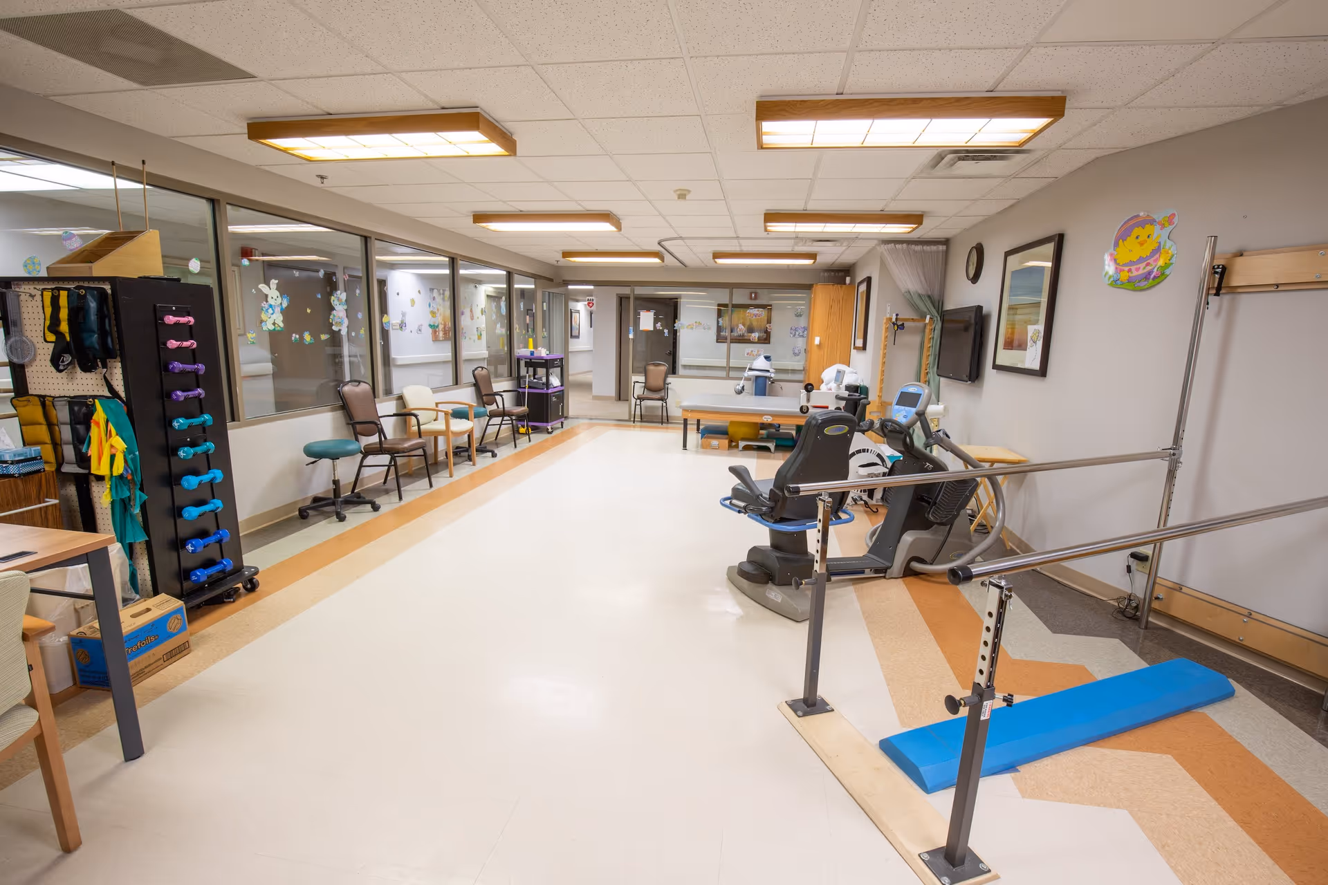 A spacious rehabilitation or physical therapy room in a healthcare facility with exercise equipment including a recumbent bike, parallel bars, and a therapy table. Several chairs are lined up against the wall with large windows showing a decorated hallway. The room is well-lit with ceiling lights and has colorful decorations on the walls.