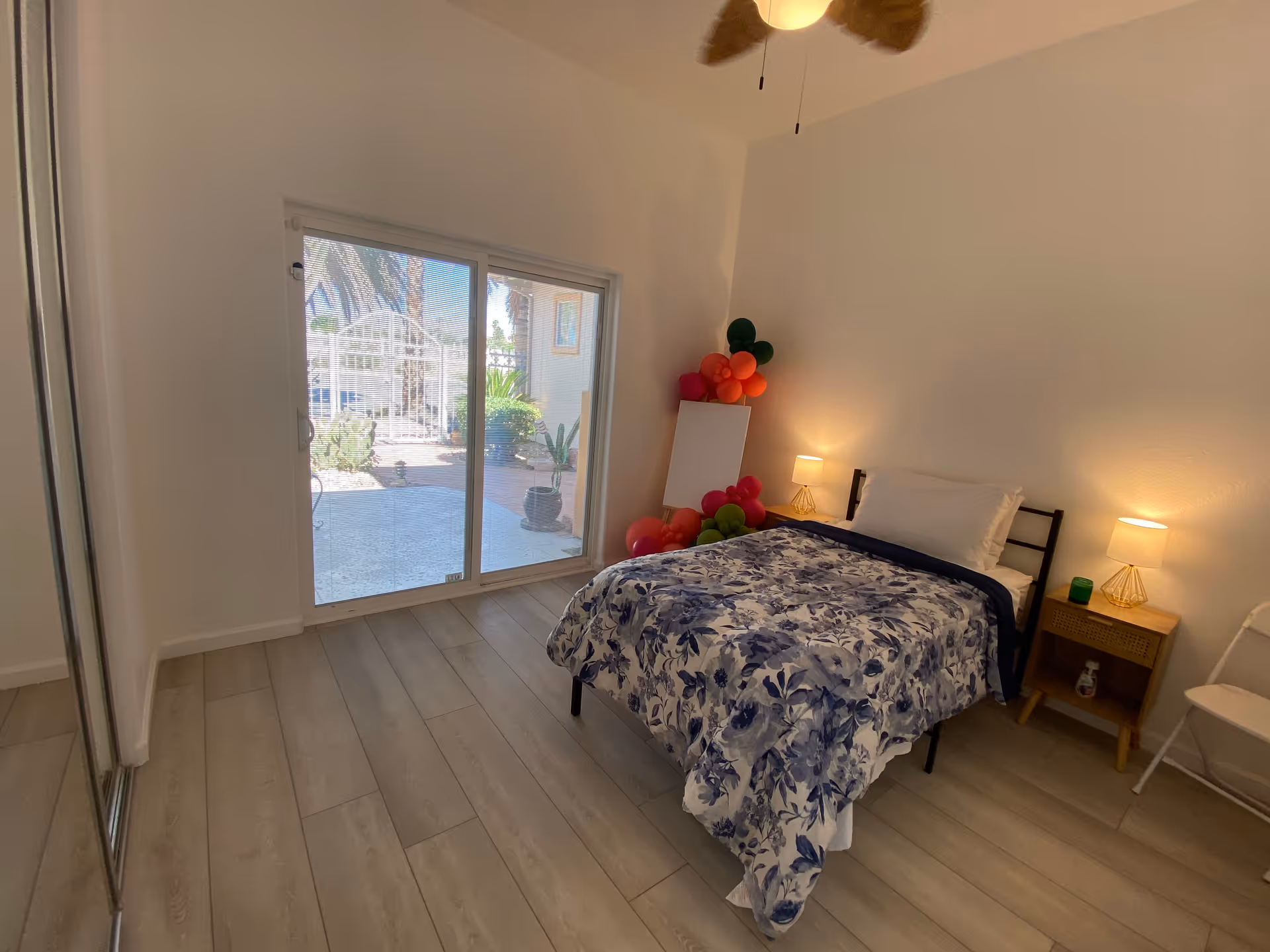 A bedroom with a single bed covered in a blue and white floral comforter, two bedside tables with lamps, a white folding chair, and a sliding glass door leading to an outdoor area with plants and a gate. There are colorful balloons and a blank canvas leaning against the wall in the corner.