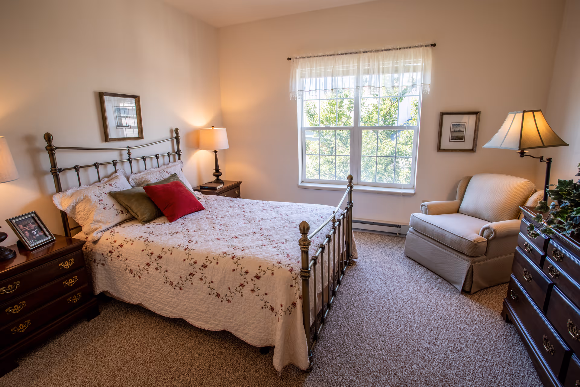 A cozy bedroom with a metal frame bed covered in a floral quilt and several pillows. There are two wooden nightstands with lamps on either side of the bed. A framed picture hangs above the bed. A large window with sheer curtains lets in natural light. A comfortable armchair and a wooden dresser with a lamp and a plant are also visible in the room.