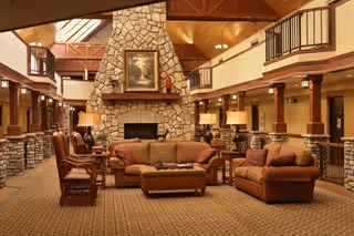 Spacious senior living facility common area with a large stone fireplace in the center, surrounded by comfortable brown sofas and chairs. The room features wooden beams on the ceiling, warm lighting from table lamps, and a second-floor balcony overlooking the area.