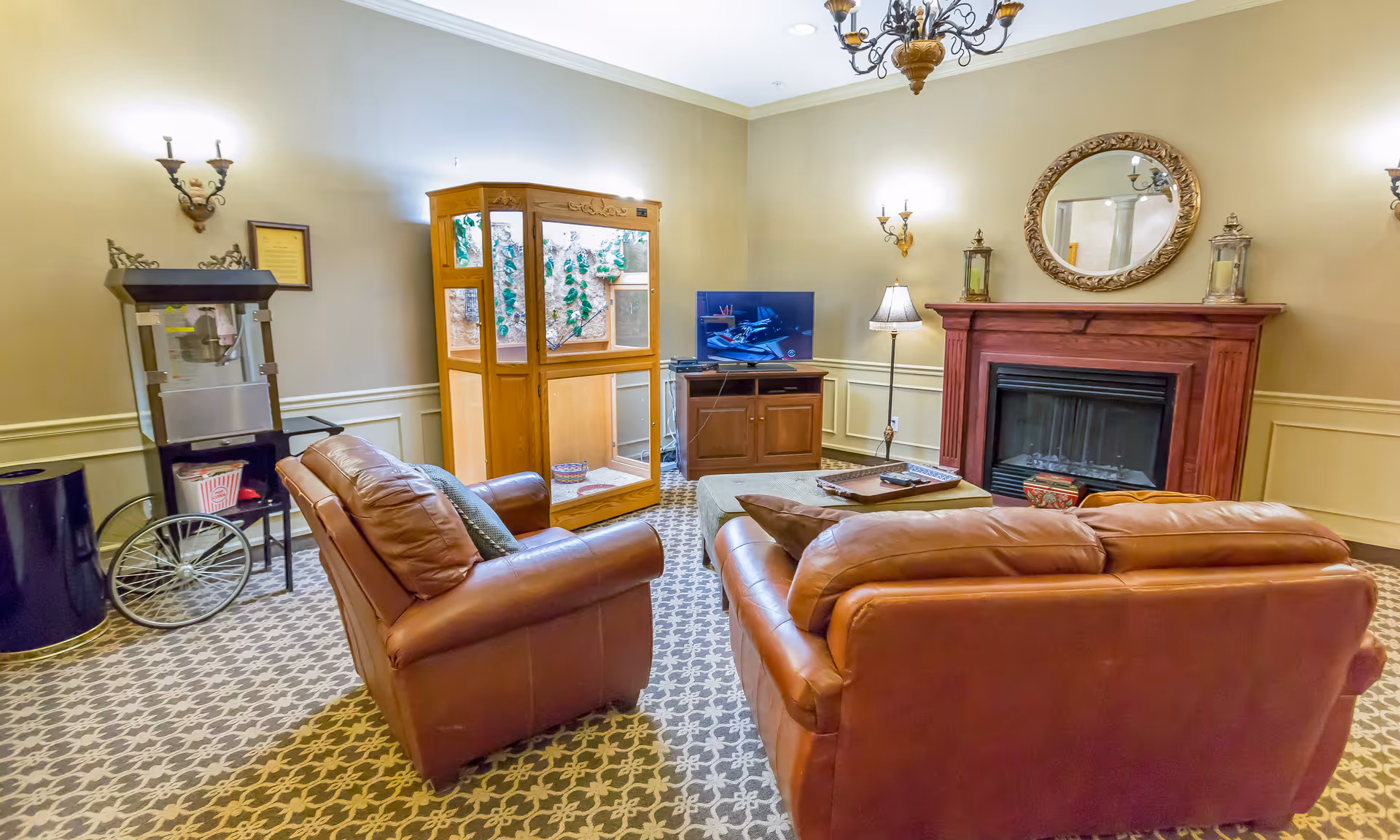 A cozy lounge with two brown leather sofas facing a TV and fireplace, with an aquarium and popcorn cart against the wall.