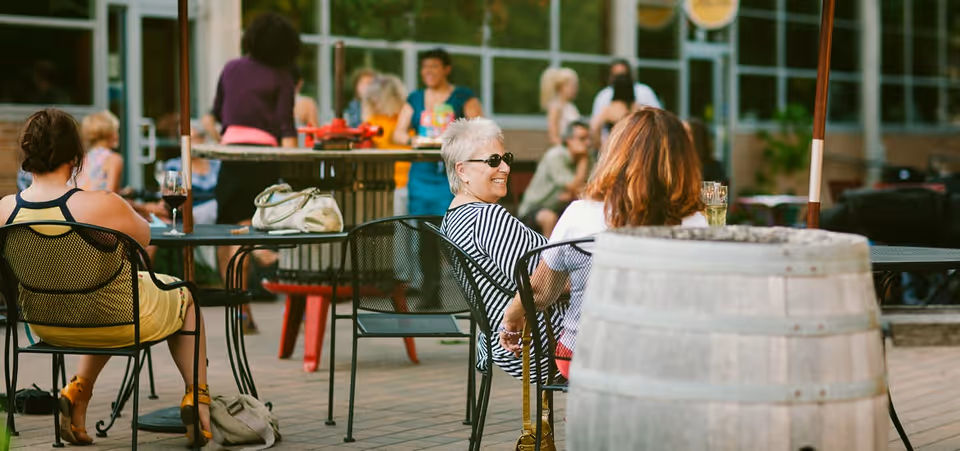 People sitting and socializing at outdoor tables on a patio with metal chairs and a wooden barrel in the foreground, surrounded by greenery and a building with large windows in the background.
