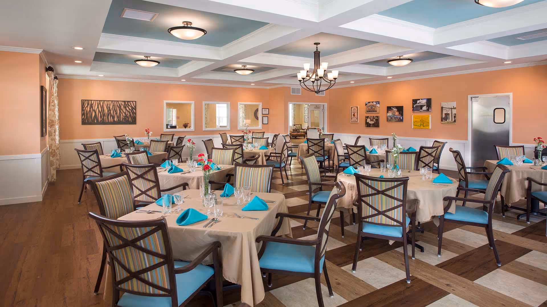 A spacious dining room with multiple round tables covered with beige tablecloths, each set with turquoise napkins, glassware, and silverware. The chairs have wooden frames with striped upholstery and turquoise seats. The room has peach-colored walls, a coffered ceiling with light blue accents, and several light fixtures including a chandelier. Decorative artwork and windows are visible on the walls.