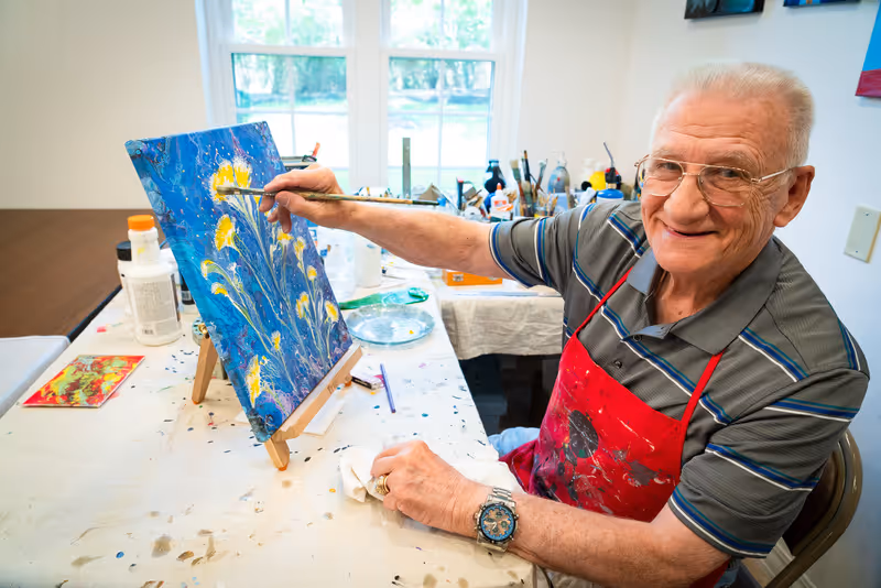 An elderly man wearing glasses and a red apron is sitting at a table painting a colorful floral artwork on a canvas. The room is well-lit with natural light coming through a window behind him, and various painting supplies are scattered on the table.