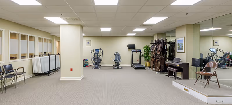 A spacious indoor room with exercise equipment including two stationary bikes and a treadmill against the far wall. The room has beige walls, a carpeted floor, and a drop ceiling with fluorescent lights. On the right side, there is a piano with a chair in front of it, a large mirror covering the wall, and a stack of folded chairs. On the left side, there are several chairs lined up against the wall and some folded tables. A potted plant and framed pictures decorate the space.