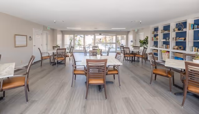 Spacious dining area with multiple tables and chairs, large windows at the far end, and shelving with books and decor on the right.