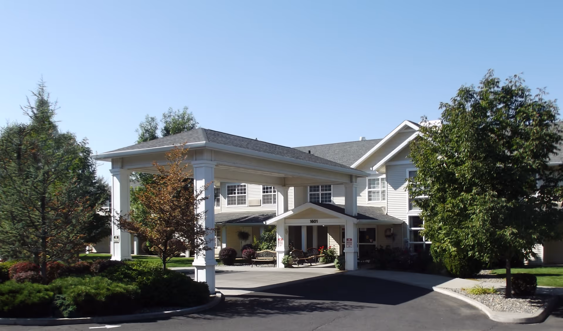 Exterior view of McKay Creek Assisted Living facility showing the entrance with a covered drop-off area, surrounded by trees and landscaping under a clear blue sky.