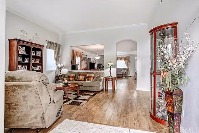 A spacious living room with wooden flooring, featuring a beige recliner chair, a sofa with patterned cushions, a wooden coffee table, and a matching side table with a lamp. There is a wooden bookshelf with framed photos on the left and a curved glass display cabinet with decorative items and a tall floral arrangement on the right. The room opens into another seating area visible through an archway.