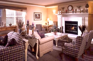 Cozy living room with plaid upholstered armchairs and a beige sofa arranged around a white coffee table. A fireplace with a mantel decorated with candles, a clock, and small plants is lit, creating a warm ambiance. The room has soft lighting from a table lamp and wall sconces, with a window covered by blinds and a wooden sideboard with decorative items.