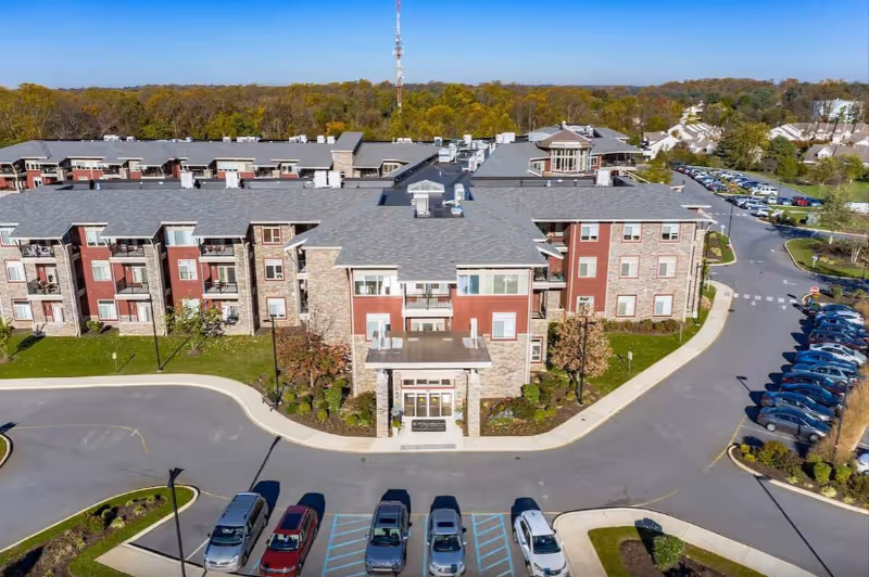 Aerial view of a large, multi-story senior living facility named The Summit, featuring a stone and red exterior with multiple windows and balconies. The building is surrounded by a parking lot with several cars parked and landscaped greenery. Trees and other residential buildings are visible in the background under a clear blue sky.