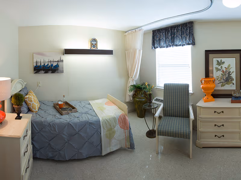 A cozy bedroom in a senior living facility with a single bed covered in a gray and patterned quilt, a wooden nightstand with a lamp and decorative items, a chair near a window with blinds and a floral valance, a small round side table, and a dresser with an orange vase and framed artwork on top.