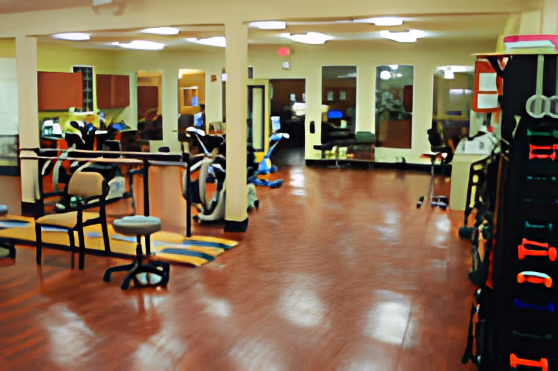 Interior view of a senior living facility's exercise or rehabilitation room with exercise equipment, chairs, and mirrors on the walls. The floor is a polished reddish-brown surface, and the room is well-lit with ceiling lights.