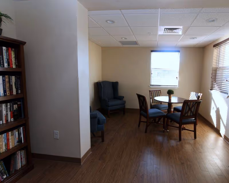 A small room with wooden flooring and beige walls featuring a round wooden table with four chairs around it. There are two windows with blinds letting in natural light. In the corner, there are two upholstered armchairs. A bookshelf filled with books is visible on the left side of the image.