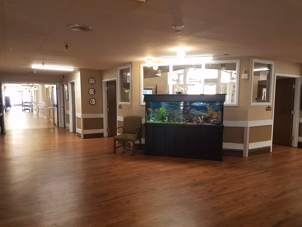 Interior hallway of a senior living facility with wood flooring, beige walls, and white trim. A large fish tank with colorful fish is placed against a wall with windows, next to a green upholstered chair. Several doors and wall decorations are visible along the hallway.