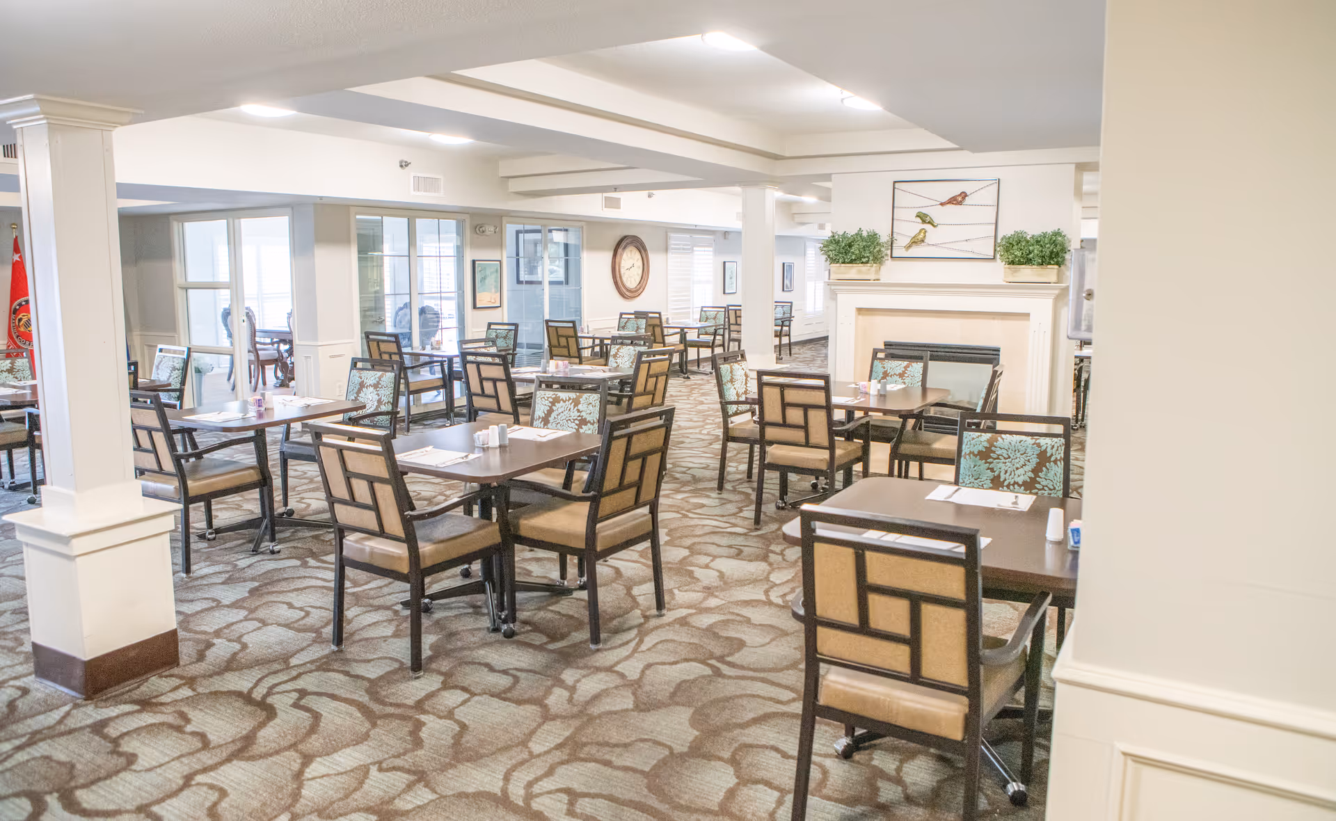 A spacious dining room with multiple square tables and cushioned chairs arranged neatly. The room has a patterned carpet, white walls, and a fireplace decorated with potted plants and a bird-themed wall art above it. Large windows allow natural light to fill the space.