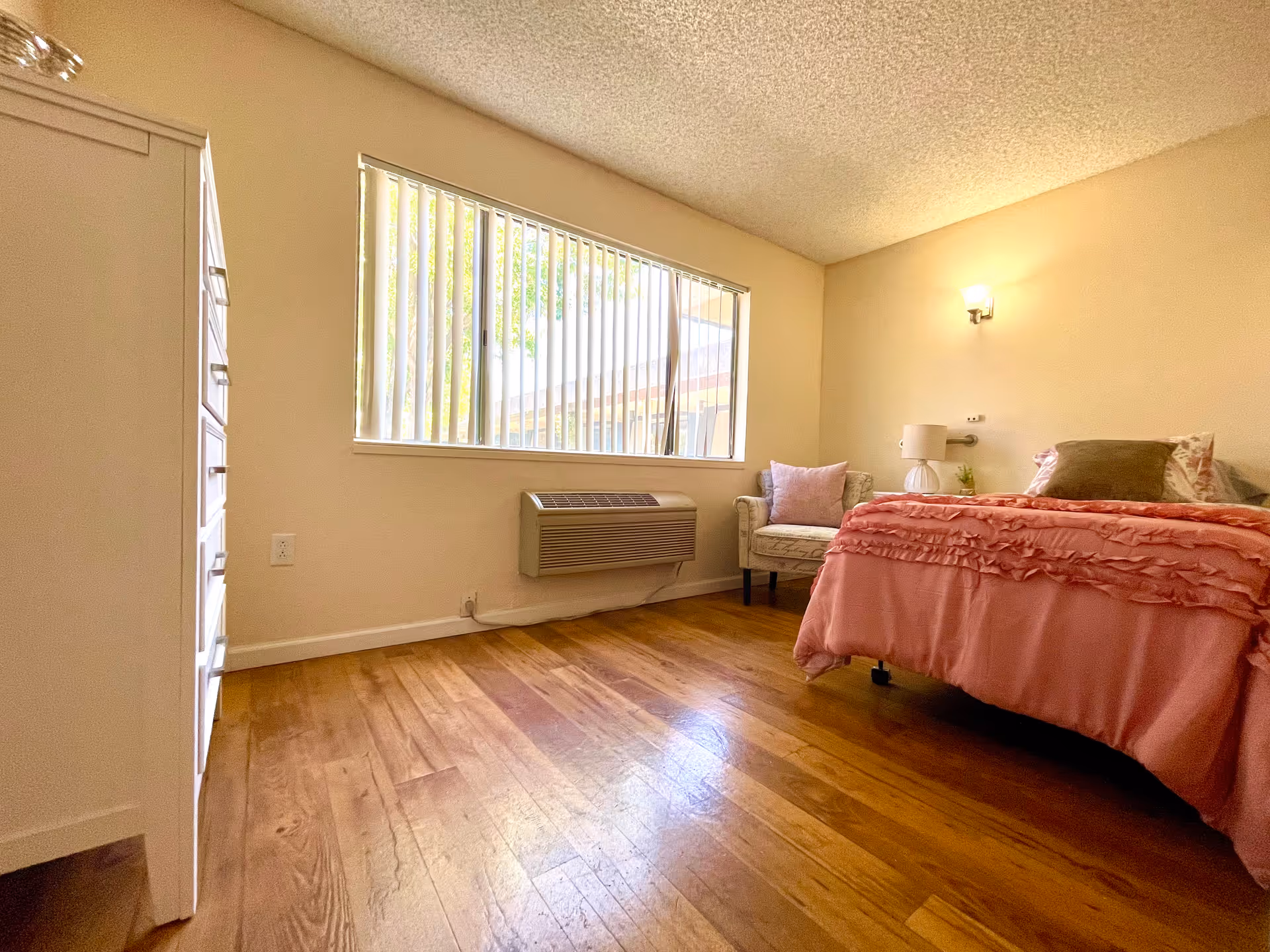 A bedroom with wooden flooring, a large window with vertical blinds, a white dresser on the left, a bed with pink bedding and pillows on the right, a small armchair with a pink pillow next to the bed, and a wall-mounted light above the bed.